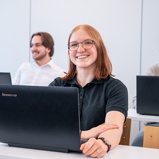 eine junge Dame arbeitet am Laptop und grinst in dei Kamera im Hintergrund ist noch ein jungerHerr zu sehen.