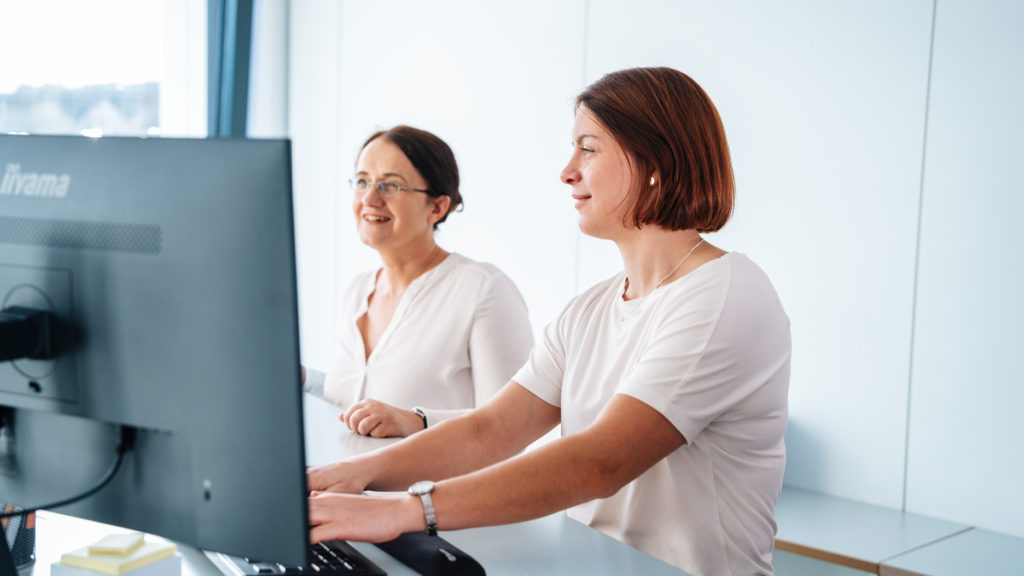 zwei Damen am Arbeitsplatz schauen in den Computer rein und sehen glücklich aus